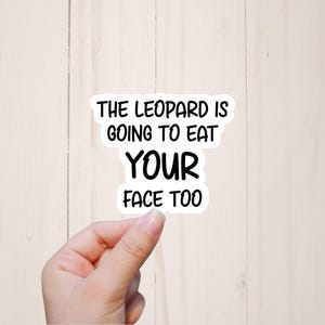 May include: A white sticker with black text that reads "THE LEOPARD IS GOING TO EAT YOUR FACE TOO". The sticker is held by a hand against a light wood grain background.