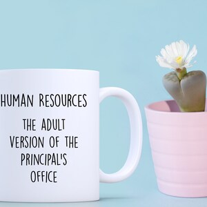 May include: White ceramic mug with the text "HUMAN RESOURCES THE ADULT VERSION OF THE PRINCIPAL'S OFFICE" in black. A small pink pot with a succulent plant and white flower sits to the right. The background is light blue.
