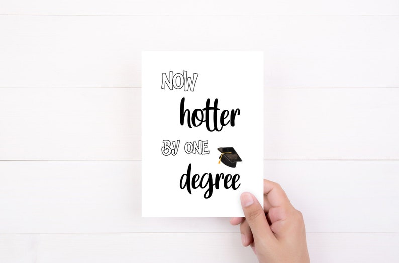 May include: A white graduation card with the text "Now hotter by one degree" in black script and a small graduation cap illustration. The card is held by a hand against a white wooden background.