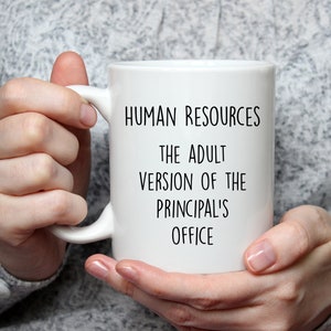 May include: White ceramic mug with the text "HUMAN RESOURCES THE ADULT VERSION OF THE PRINCIPAL'S OFFICE" in black. The mug is held by a person wearing a gray sweater.