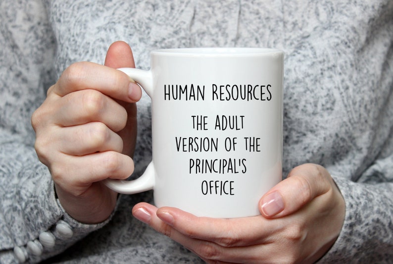 May include: White ceramic mug with the text "HUMAN RESOURCES THE ADULT VERSION OF THE PRINCIPAL'S OFFICE" in black. The mug is held by a person wearing a gray sweater.