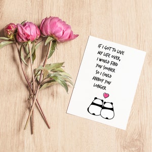 Op de afbeelding: Een witte wenskaart met de tekst "IF I GOT TO LIVE MY LIFE OVER, I WOULD FIND YOU SOONER SO I COULD ANNOY YOU LONGER" en panda-illustraties. Roze pioenrozen en knoppen zijn gerangschikt op een licht houten oppervlak.