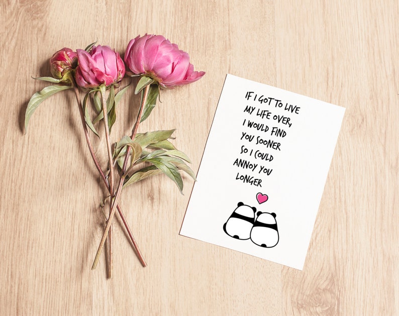 Op de afbeelding: Een witte wenskaart met de tekst "IF I GOT TO LIVE MY LIFE OVER, I WOULD FIND YOU SOONER SO I COULD ANNOY YOU LONGER" en panda-illustraties. Roze pioenrozen en knoppen zijn gerangschikt op een licht houten oppervlak.