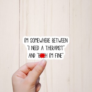 May include: White sticker with black text that reads "I'm somewhere between 'I need a therapist' and 'b*tch I'm fine'".