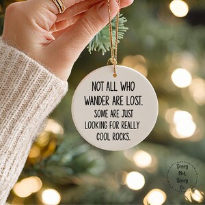 Puede incluir: Un adorno navide&ntilde;o de cer&aacute;mica blanca con una cuerda dorada, con el texto "NOT ALL WHO WANDER ARE LOST. SOME ARE JUST LOOKING FOR REALLY COOL ROCKS." El adorno se sostiene frente a un &aacute;rbol de Navidad.