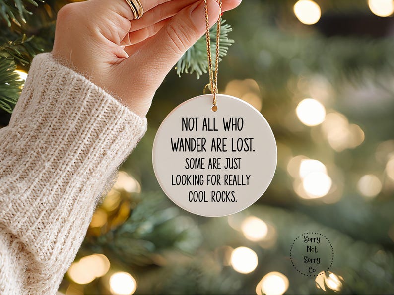 Puede incluir: Un adorno navide&ntilde;o de cer&aacute;mica blanca con una cuerda dorada, con el texto "NOT ALL WHO WANDER ARE LOST. SOME ARE JUST LOOKING FOR REALLY COOL ROCKS." El adorno se sostiene frente a un &aacute;rbol de Navidad.