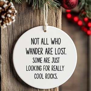 Puede incluir: Adorno redondo de cer&aacute;mica blanca con el texto "NOT ALL WHO WANDER ARE LOST. SOME ARE JUST LOOKING FOR REALLY COOL ROCKS." Cuelga de una cuerda, con fondo de madera y adornos navide&ntilde;os.