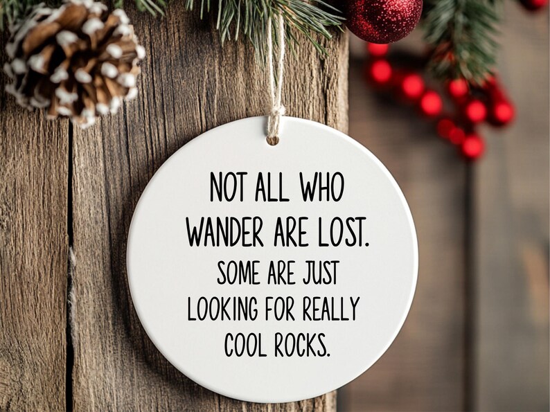Puede incluir: Adorno redondo de cer&aacute;mica blanca con el texto "NOT ALL WHO WANDER ARE LOST. SOME ARE JUST LOOKING FOR REALLY COOL ROCKS." Cuelga de una cuerda, con fondo de madera y adornos navide&ntilde;os.