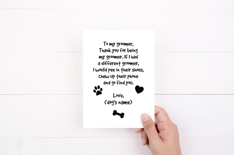May include: A white greeting card with black text and paw print, heart, and bone graphics. The text reads: "To my groomer, Thank you for being my groomer...Love, (dog's name)". The card is held by a person.