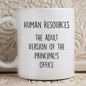 May include: White ceramic mug with a black handle. The mug features the text "HUMAN RESOURCES THE ADULT VERSION OF THE PRINCIPAL'S OFFICE" in black, centered on the front. The mug is on a white surface.