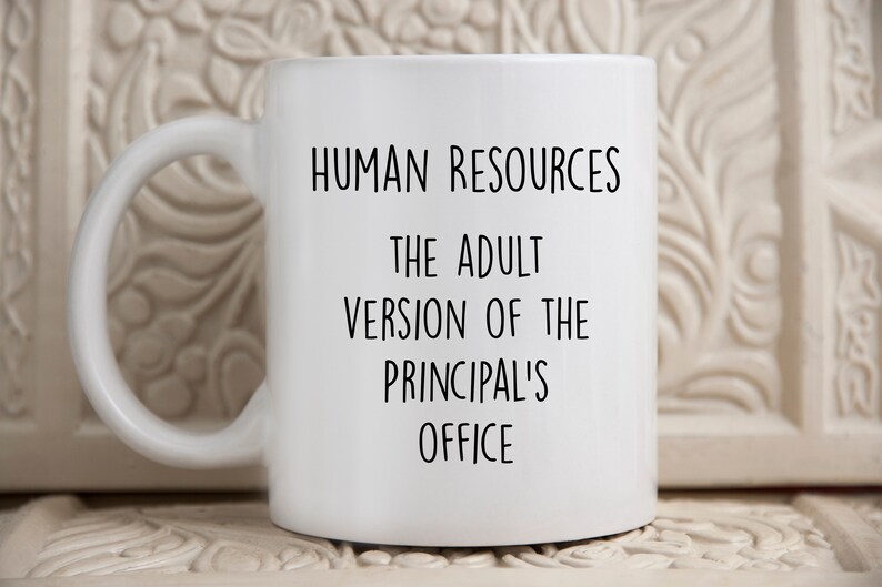 May include: White ceramic mug with a black handle. The mug features the text "HUMAN RESOURCES THE ADULT VERSION OF THE PRINCIPAL'S OFFICE" in black, centered on the front. The mug is on a white surface.