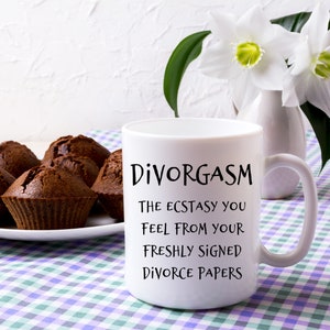 Puede incluir: Taza de caf&eacute; blanca con texto negro que dice "Divorgasm The Ecstasy You Feel From Your Freshly Signed Divorce Papers". La taza est&aacute; sobre un mantel a cuadros con magdalenas de chocolate en un plato blanco en el fondo.