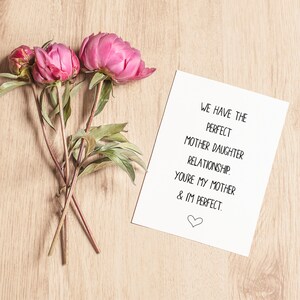 May include: A white card with the text "WE HAVE THE PERFECT MOTHER DAUGHTER RELATIONSHIP. YOU'RE MY MOTHER & IM PERFECT." with a small heart symbol at the bottom. Three pink peonies are on a wooden surface next to the card.