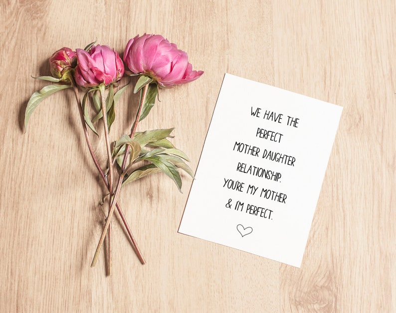 May include: A white card with the text "WE HAVE THE PERFECT MOTHER DAUGHTER RELATIONSHIP. YOU'RE MY MOTHER & IM PERFECT." with a small heart symbol at the bottom. Three pink peonies are on a wooden surface next to the card.