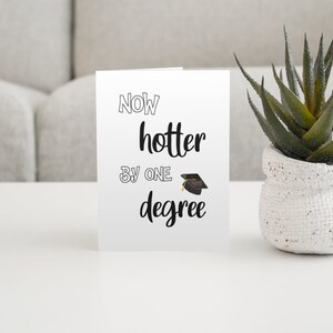 May include: A white graduation card with the text "Now hotter by one degree" and a graduation cap illustration. A small potted succulent plant sits to the right of the card on a white surface.