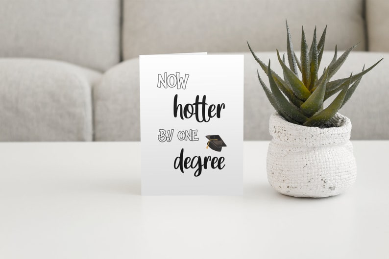 May include: A white graduation card with the text "Now hotter by one degree" and a graduation cap illustration. A small potted succulent plant sits to the right of the card on a white surface.