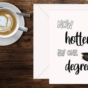 May include: A greeting card with the text "Now hotter by one degree" and a graduation cap illustration. A cup of coffee with latte art sits on a saucer to the left of the card. The background is a dark wood surface.