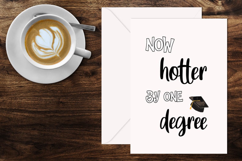 May include: A greeting card with the text "Now hotter by one degree" and a graduation cap illustration. A cup of coffee with latte art sits on a saucer to the left of the card. The background is a dark wood surface.