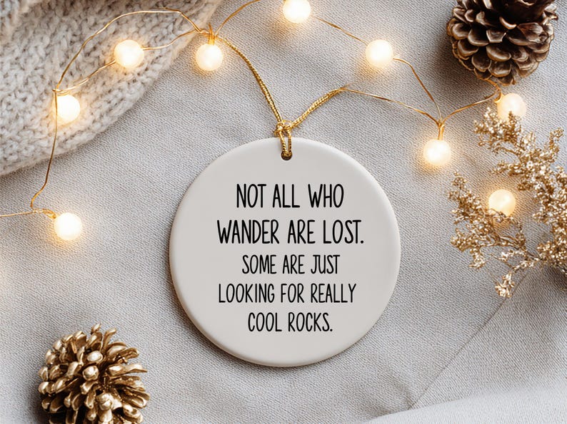 Puede incluir: Adorno redondo de cer&aacute;mica blanca con el texto "NOT ALL WHO WANDER ARE LOST. SOME ARE JUST LOOKING FOR REALLY COOL ROCKS.". Se muestra colgado con una cuerda dorada. El fondo incluye luces de hadas, pi&ntilde;as y ramas decorativas doradas.