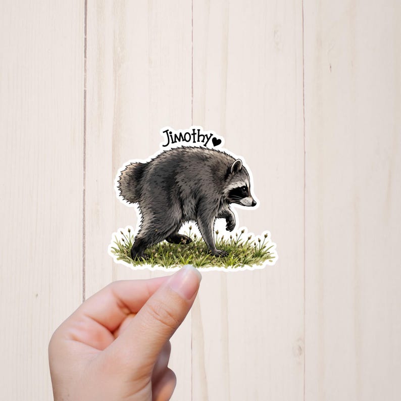 May include: A die-cut sticker featuring a gray and black raccoon walking through green grass. The raccoon is depicted in profile, facing right, with the name Jimothy and a small black heart printed in black text above its back.