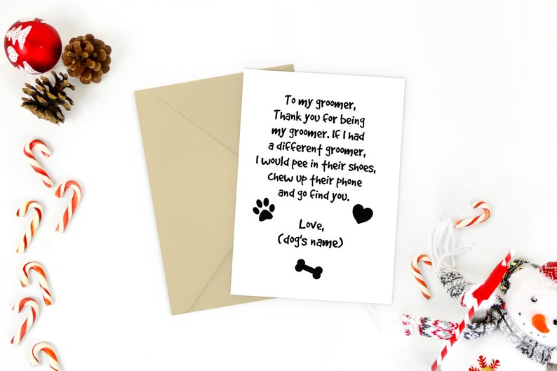 May include: A greeting card with a humorous message for a dog groomer, accompanied by a tan envelope. The card features a paw print, heart, and bone graphic. Christmas decorations, including candy canes and ornaments, surround the card.