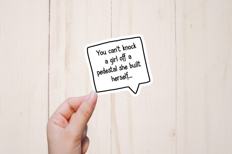 Puede incluir: Pegatina blanca con contorno negro en forma de bocadillo. El texto dentro de la pegatina dice "You can't knock a girl off a pedestal she built herself..."