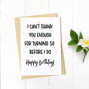 Może przedstawiać: Kartka urodzinowa z napisem "I can't thank you enough for turning 50 before I do. Happy Birthday!". Kartka leży na białej powierzchni, wraz z kopertą z papieru kraft i gałązką kwiatową.
