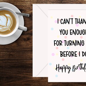 Może przedstawiać: Karta urodzinowa z napisem "I can't thank you enough for turning 50 before I do. Happy Birthday!" jest wyświetlana obok białej koperty. Filiżanka kawy z latte art stoi na spodku z łyżeczką.