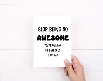 May include: A white greeting card with black text that reads "STOP BEING SO AWESOME. YOU'RE MAKING THE REST OF US LOOK BAD." The card is held by a hand against a white wood-grain background.