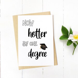 May include: A graduation card with the text "Now hotter by one degree" in black script and a mortarboard illustration. The card is on a kraft paper envelope, with a white flower and green leaves on a white wooden surface.