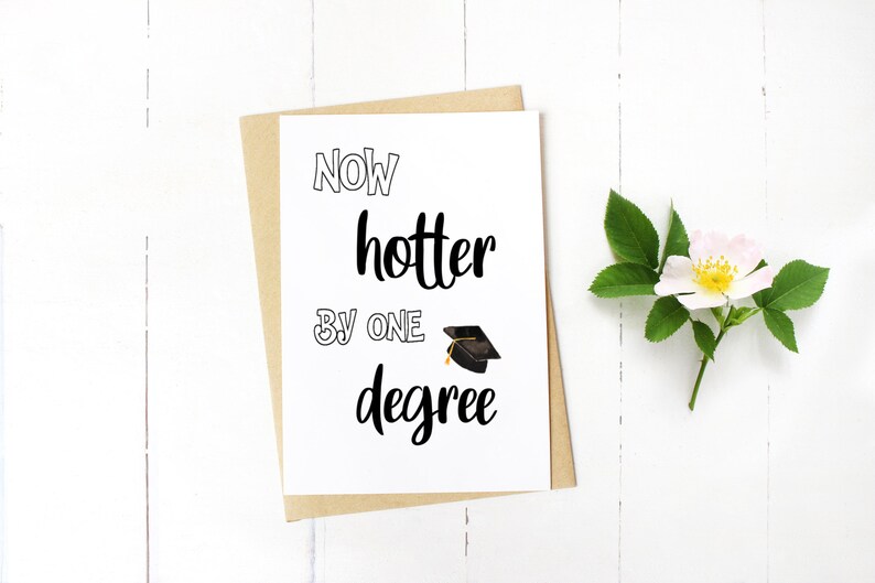 May include: A graduation card with the text "Now hotter by one degree" in black script and a mortarboard illustration. The card is on a kraft paper envelope, with a white flower and green leaves on a white wooden surface.