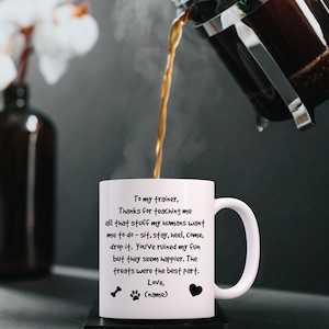 Puede incluir: Taza de caf&eacute; de cer&aacute;mica blanca con una huella de pata negra y un dise&ntilde;o de hueso. La taza tiene un mensaje que dice: "A mi entrenador, Gracias por ense&ntilde;arme todo eso que mis humanos quieren que haga - sentarme, quedarme, tal&oacute;n, venir, soltar. Has arruinado mi diversi&oacute;n, pero ellos parecen m&aacute;s felices. Las golosinas fueron la mejor parte. Amor, (nombre)"