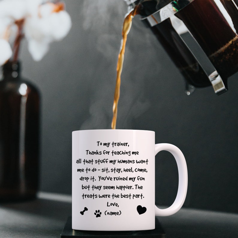 Puede incluir: Taza de caf&eacute; de cer&aacute;mica blanca con una huella de pata negra y un dise&ntilde;o de hueso. La taza tiene un mensaje que dice: "A mi entrenador, Gracias por ense&ntilde;arme todo eso que mis humanos quieren que haga - sentarme, quedarme, tal&oacute;n, venir, soltar. Has arruinado mi diversi&oacute;n, pero ellos parecen m&aacute;s felices. Las golosinas fueron la mejor parte. Amor, (nombre)"