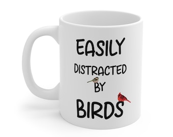 May include: A white ceramic mug featuring the black text EASILY DISTRACTED BY BIRDS. The design includes a small brown and white bird perched on the letter Y and a bright red cardinal bird resting on the letter S in the word BIRDS.