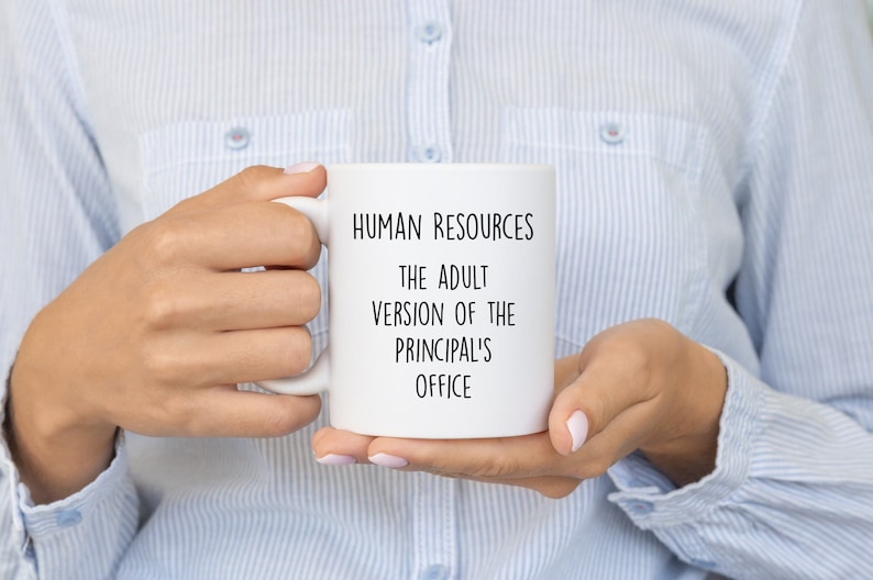 May include: White ceramic mug with the text "HUMAN RESOURCES THE ADULT VERSION OF THE PRINCIPAL'S OFFICE" in black. The mug is held by a person wearing a light blue button-down shirt.