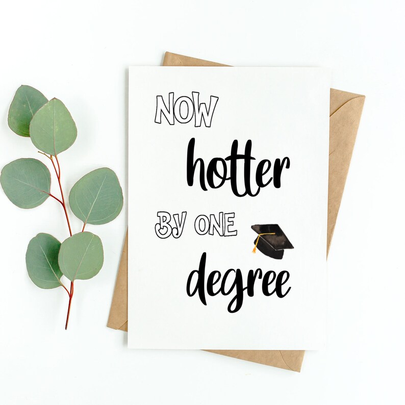 May include: A white graduation card with the text "NOW hotter BY ONE degree" in black script. A small graduation cap illustration is next to the word "degree". The card is inside a brown envelope, with eucalyptus leaves in the background.