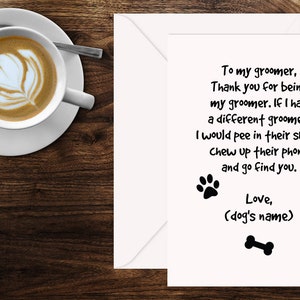 May include: A greeting card with a white envelope, featuring a humorous message for a dog groomer. The card includes text, paw prints, a heart, and a bone graphic. A cup of coffee with latte art sits on a saucer to the left.