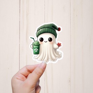 Puede incluir: Un lindo fantasma de dibujos animados con un sombrero verde y sosteniendo un batido verde con una pajita. El fantasma sonr&iacute;e y tiene una flor roja en su mano.