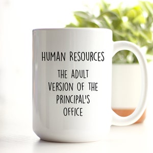 May include: White ceramic mug with a large handle. The mug has black text that reads "HUMAN RESOURCES THE ADULT VERSION OF THE PRINCIPAL'S OFFICE". The mug is in front of a blurred background with green foliage.