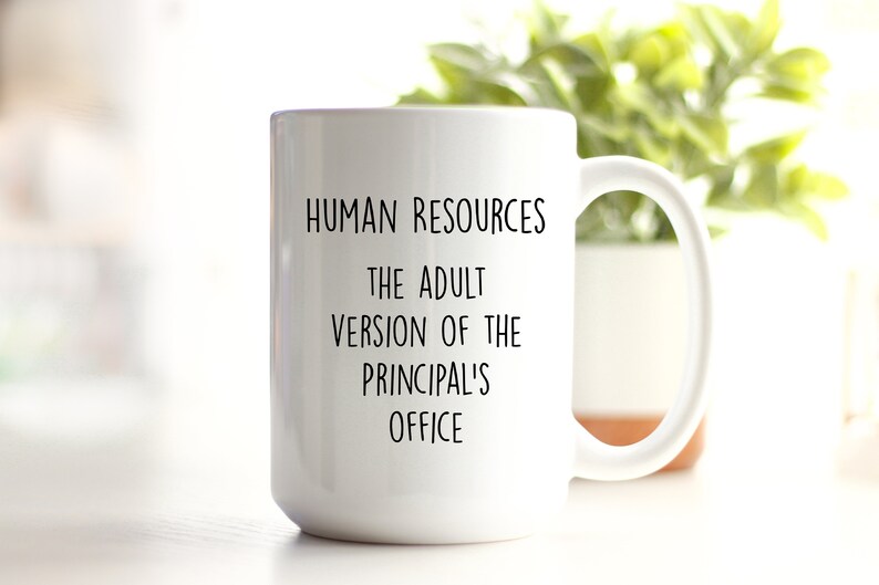 May include: White ceramic mug with a large handle. The mug has black text that reads "HUMAN RESOURCES THE ADULT VERSION OF THE PRINCIPAL'S OFFICE". The mug is in front of a blurred background with green foliage.