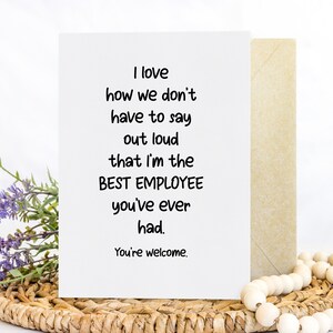 May include: A white greeting card with black text that reads: I love how we don't have to say out loud that I'm the BEST EMPLOYEE you've ever had. You're welcome. The card is positioned next to a tan envelope, a sprig of purple flowers, and wooden beads.