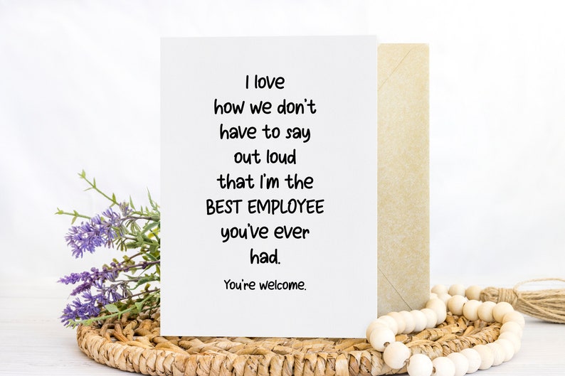 May include: A white greeting card with black text that reads: I love how we don't have to say out loud that I'm the BEST EMPLOYEE you've ever had. You're welcome. The card is positioned next to a tan envelope, a sprig of purple flowers, and wooden beads.