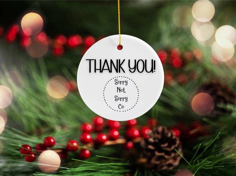 Puede incluir: Adorno redondo de cer&aacute;mica blanca con las palabras "THANK YOU!" en negro. Debajo, un c&iacute;rculo punteado contiene el texto "Sorry Not Sorry Co". El adorno cuelga de una cuerda dorada sobre un fondo festivo de vegetaci&oacute;n, bayas rojas y luces bokeh.