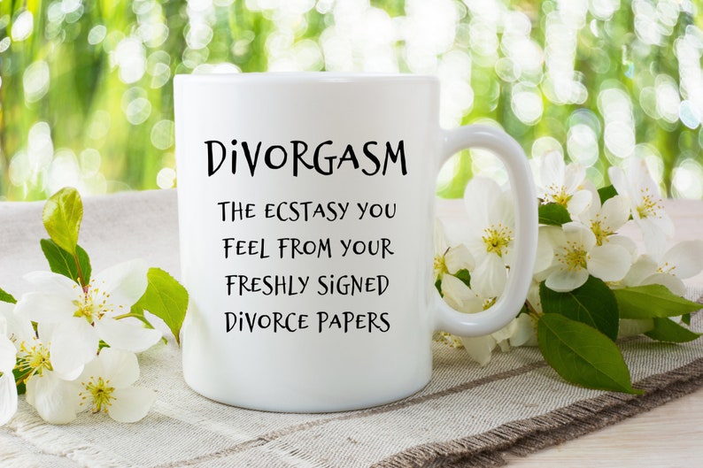 Puede incluir: Taza de caf&eacute; de cer&aacute;mica blanca con texto negro que dice "Divorgasm: The ecstasy you feel from your freshly signed divorce papers". La taza est&aacute; sobre una superficie de lino blanco con flores blancas.