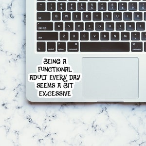 Pu&ograve; includere: Un computer portatile bianco con una tastiera nera e un adesivo sulla parte superiore del touchpad che recita "Being a functional adult every day seems a bit excessive". Il laptop &egrave; su una superficie di marmo bianco.