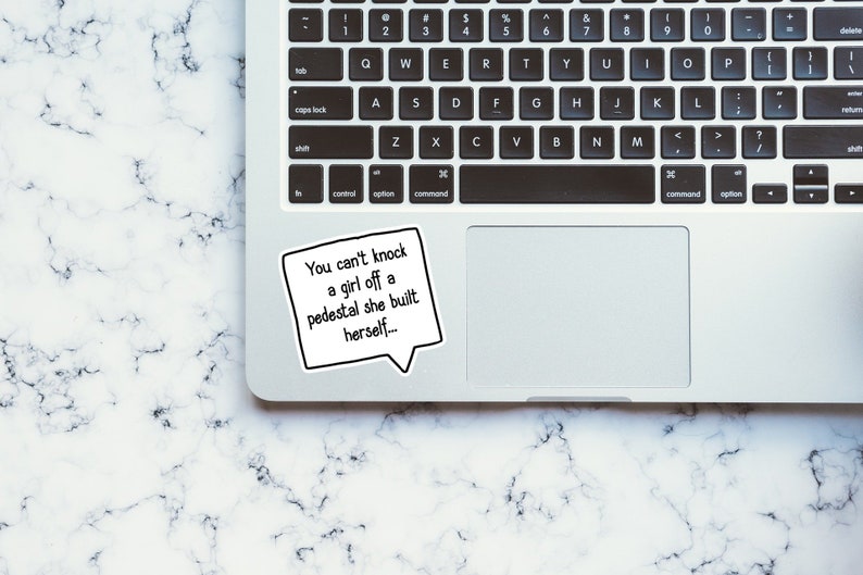 Puede incluir: Un port&aacute;til blanco con un teclado negro y un trackpad negro. Una pegatina con el texto "You can't knock a girl off a pedestal she built herself..." est&aacute; en el port&aacute;til.