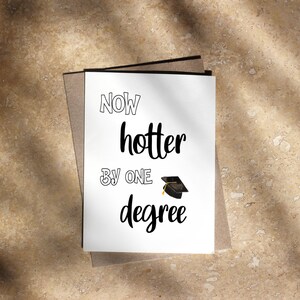 May include: A graduation card with the words "Now hotter by one degree" in black script. A small graduation cap illustration is below the text. The card is white and set against a neutral background.