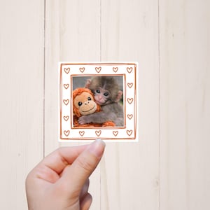 May include: A square sticker featuring a photograph of a baby monkey hugging an orange stuffed animal. The photo is framed with a brown border decorated with small hearts. The sticker is held by a hand.