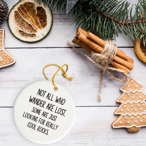 Puede incluir: Una composici&oacute;n festiva con un adorno de cer&aacute;mica blanca con el texto "NOT ALL WHO WANDER ARE LOST. SOME ARE JUST LOOKING FOR REALLY COOL ROCKS." y galletas de jengibre. Tambi&eacute;n hay palitos de canela y rodajas de naranja secas.