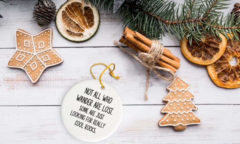 Puede incluir: Una composici&oacute;n festiva con un adorno de cer&aacute;mica blanca con el texto "NOT ALL WHO WANDER ARE LOST. SOME ARE JUST LOOKING FOR REALLY COOL ROCKS." y galletas de jengibre. Tambi&eacute;n hay palitos de canela y rodajas de naranja secas.